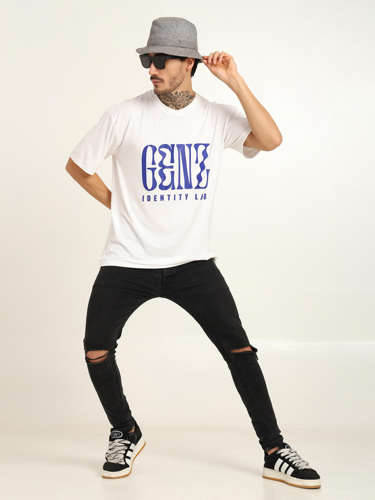 Man in oversized white bamboo t-shirt with blue graphic design, stylishly posing in black ripped jeans and hat.