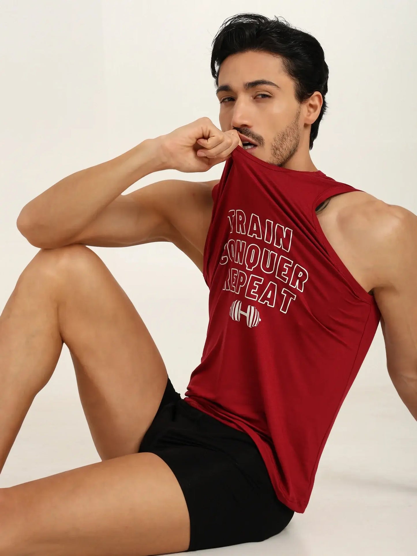 Man in red tank top with text 'Train Conquer Repeat' sitting in black trunks, showcasing a fitness lifestyle.
